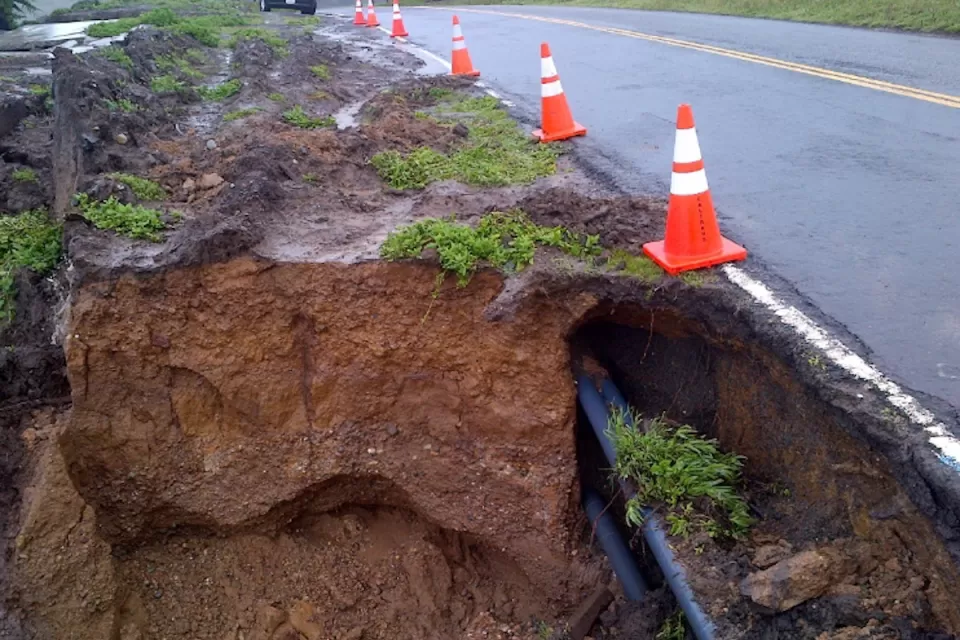 Eroded cliffside and exposed utilities along asphalt Highway 1 in California with orange traffic cones lining side of road.